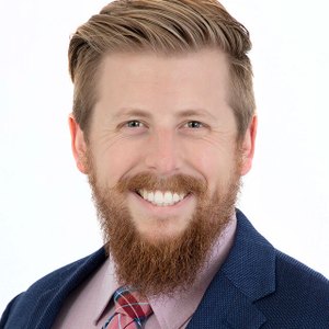 Headshot of Patrick Reese who has combed brown hair, a beard, and a blue suit with pink shirt and colorful tie